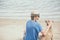 Young handsome man wearing blue t-shirt, hat and sunglasses, sitting on the beach with the dog in Thailand