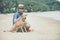 Young handsome man wearing blue t-shirt, hat and sunglasses, sitting on the beach with the dog in Thailand