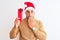 Young handsome man wearing christmas hat and holding gift over isolated background cover mouth with hand shocked with shame for