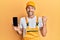 Young handsome man wearing handyman uniform showing smartphone screen screaming proud, celebrating victory and success very