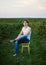 Young handsome man, wearing torn jeans and white t-shirt, sitting on yellow stool on green field, thinking. Artistic education