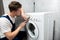 Young handsome man worker in uniform repairing washing machine at home in the toilette professional repair service