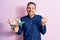 Young handsome romantic man holding beautiful bouquet of flowers over pink background screaming proud, celebrating victory and