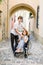 Young handsome smiling man and his pretty wife in wheelchair posing together on the background of old city center, arch