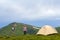 Young handsome smiling man standing on green grassy valley on sunny day at small tourist tent mountains and bright blue sky backgr