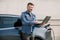 Young handsome smiling man working on laptop, waiting while his luxury electric car is charching, paying for charging