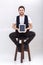 Young handsome smilling man with beard in white shirt and black waistcoat holding tablet and sits on chair on gray