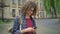 Young handsome student with curly hair in glasses texting on phone and holding backpack, standing in park near