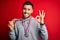 Young handsome succesful man winning medals standing over isolated red background doing ok sign with fingers, excellent symbol