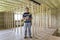 Young handsome worker with level and screwdriver looking in camera in big light attic room with oak floor, insulated ceiling and w