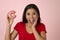 Young happy attractive Latin woman in red top smiling excited holding sugar donut on pink background