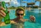 Young happy and attractive man listening to music with headset at tropical resort swimming pool drinking beer enjoying indulgent