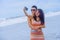 young happy and beautiful Asian Chinese couple taking selfie photo with mobile phone camera smiling joyful having fun on the beach