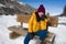 Young happy and beautiful Asian Chinese woman in yellow jacket and winter hat smiling cheerful on bech over frozen lake covered in