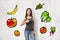 Young happy brunette girl in casual clothes showing thumb up with cartoon fruit and vegetables on white background