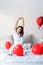 Young happy brunette woman sitting awake in the bed with red heart shaped balloons