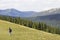 Young happy child boy with backpack walking in mountain grassy valley on background of summer woody mountain. Active lifestyle,