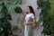 Young happy female gardener standing against lush Monstera Deliciosa houseplants in home garden