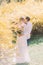Young happy groom gently embracing back of his beautiful bride in white dress outdoors with yellow flowers on background