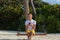 Young happy man seated on a swing and using his phone. White sand and jungle as background