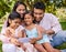 Young happy mixed race family using a phone and sitting on grass together in a park. Loving hispanic parents watching