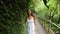 Young Happy Mixed Race Girl in White Dress Walking in Jungle Forest, Looking Around and Touching with Hand Tropical Leaves. Calm