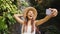 Young Happy Mixed Race Tourist Girl in White Dress and Straw Hat Making Selfie Photos Using Mobile Phone with Amazing Wild Jungle
