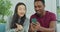 Young happy multinational couple sit on sofa use smartphone and credit card make order, buying on internet laughing