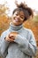 Young happy smiling mixed-race woman with coffee cup in autumn nature, pleased african american female with curly hair in knitted