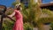 Young happy woman enjoys the sun standing in the hotel garden with palm trees wearing red dress, hat and sunglasses