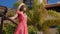 Young happy woman enjoys the sun standing in the hotel garden with palm trees wearing red dress, hat and sunglasses. Slow motion