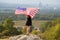 Young happy woman with long hair raising up waving on wind american national flag in her hands standing on high rocky hill