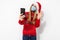 Young happy woman in a red Santa Claus hat and a medical mask on her face, waving, making a video call on a smartphone on