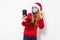 Young happy woman in a red Santa Claus hat and a medical mask on her face, waving, making a video call on a smartphone on