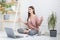 A young happy woman sits on the floor in a bright apartment or office interior and works at a laptop, freelancer girl at
