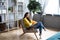 Young happy woman in a yellow sweater and jeans sitting on a cozy armchair in the living room at home