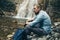 Young Hiker Man With Backpack, Sitting On The Rocks Near Waterfall Trek Hiking Destination Experience Lifestyle Concept