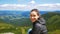 Young hiker woman smiling and looking at the camera sitting on top of a cliff on the background of mountain scenery. A