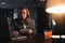 Young hipster businessman sits by the wooden table with lamp, laptop and smartphone at night office. Man with long hair work with