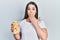 Young hispanic girl holding bottle with boiled potatoes covering mouth with hand, shocked and afraid for mistake