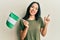 Young hispanic girl holding nigeria flag smiling happy pointing with hand and finger to the side