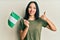 Young hispanic girl holding nigeria flag smiling happy and positive, thumb up doing excellent and approval sign