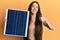 Young hispanic girl holding photovoltaic solar panel smiling happy and positive, thumb up doing excellent and approval sign