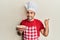 Young hispanic man chef cooking carrot cake pointing thumb up to the side smiling happy with open mouth