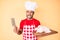 Young hispanic man cooking chicken holding knife sticking tongue out happy with funny expression