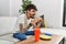 Young hispanic man eating classical burger and drinking soda sitting on the sofa at home