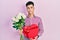 Young hispanic man holding anniversary present and bouquet of flowers relaxed with serious expression on face