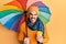 Young hispanic man holding colorful umbrella drinking take away coffee sticking tongue out happy with funny expression