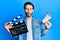 Young hispanic man holding video film clapboard and megaphone making fish face with mouth and squinting eyes, crazy and comical