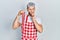 Young hispanic man with modern dyed hair wearing apron holding raw beef steak serious face thinking about question with hand on